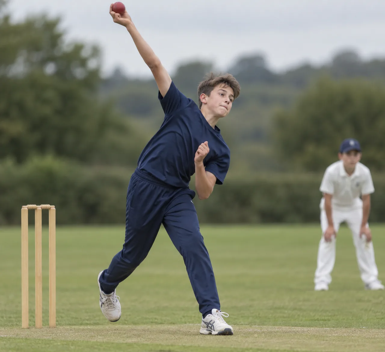 A boy runs in to bowl, bowling arm high and the red ball still in his hand, a fielder waiting in the background.